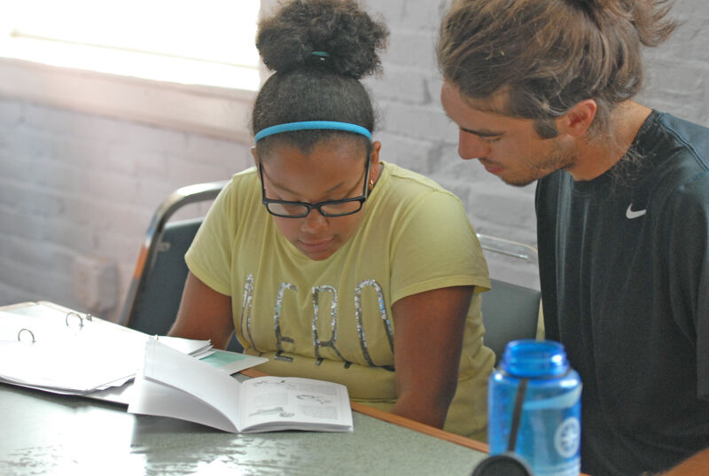 In the image, a young woman with glasses and a blue headband is intently reading a book. A man with long hair is looking over her shoulder, seemingly helping or observing. They are seated at a table, and there's a water bottle nearby. The setting appears to be indoors, possibly a classroom or study area.